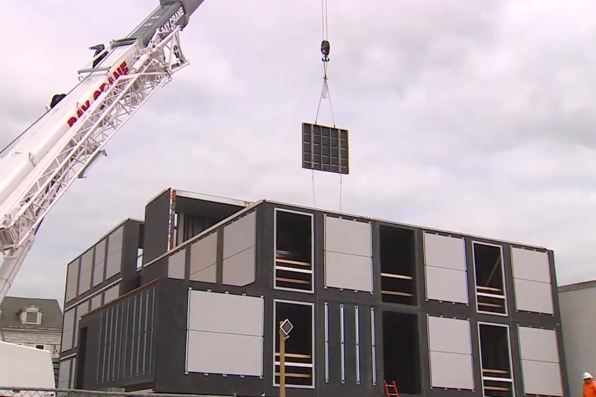 A wall panel lowered by crane onto a partially assembled Vessel building, a worker guiding it in